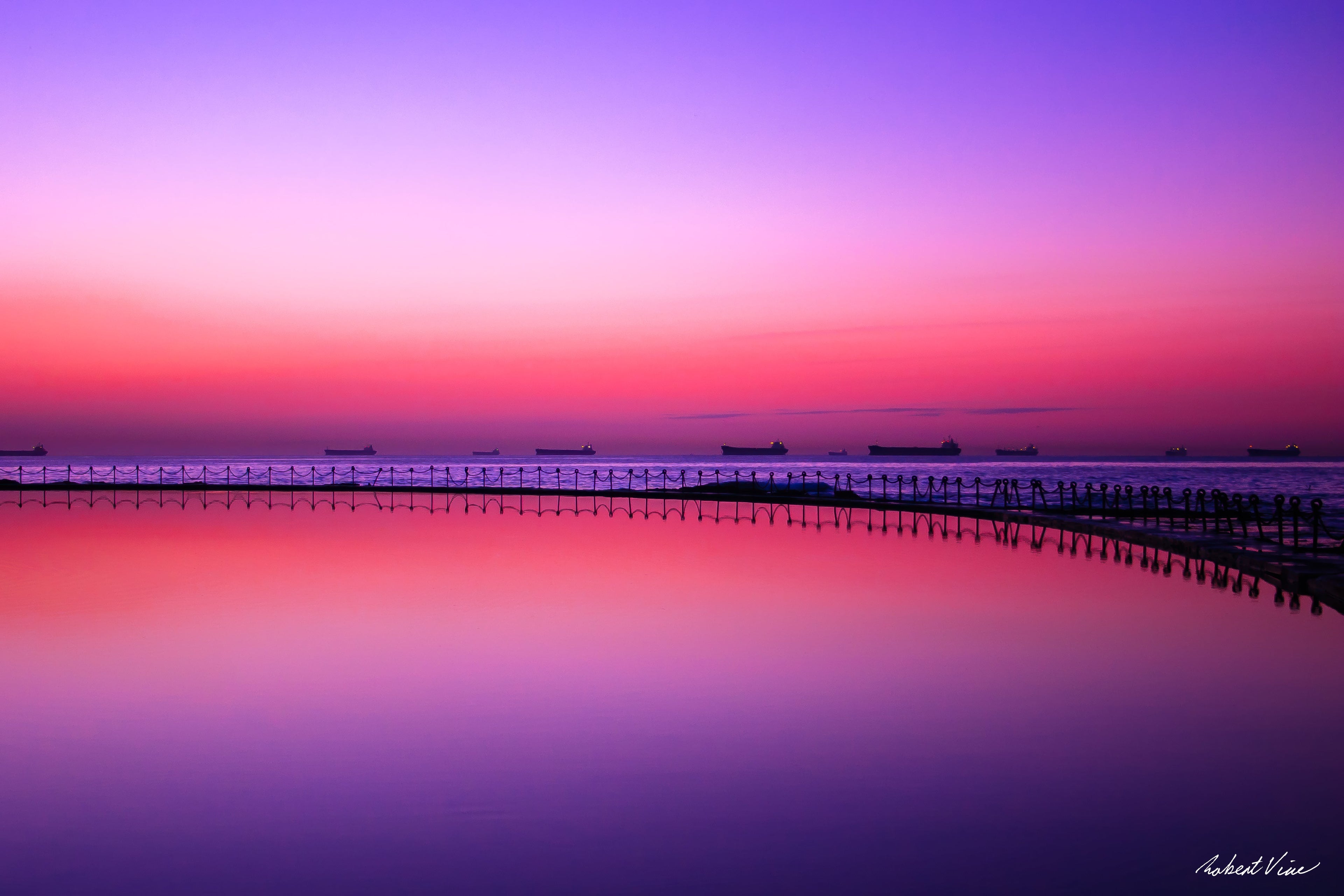 Newcastle Canoe Pool at dawn with curved walkway reflected in still water, pink and purple sky and ships on the horizon, landscape photograph by Robert Vine.
