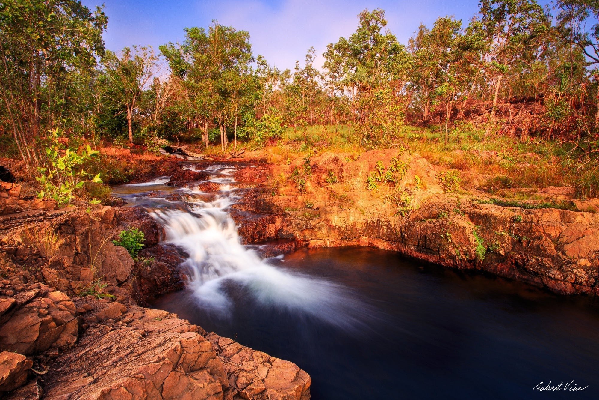 Golden morning light illuminates the cascading waters of Buley Rockhole in Litchfield National Park, Northern Territory, Australia, flowing over ancient sandstone into a tranquil swimming hole surrounded by native bushland.