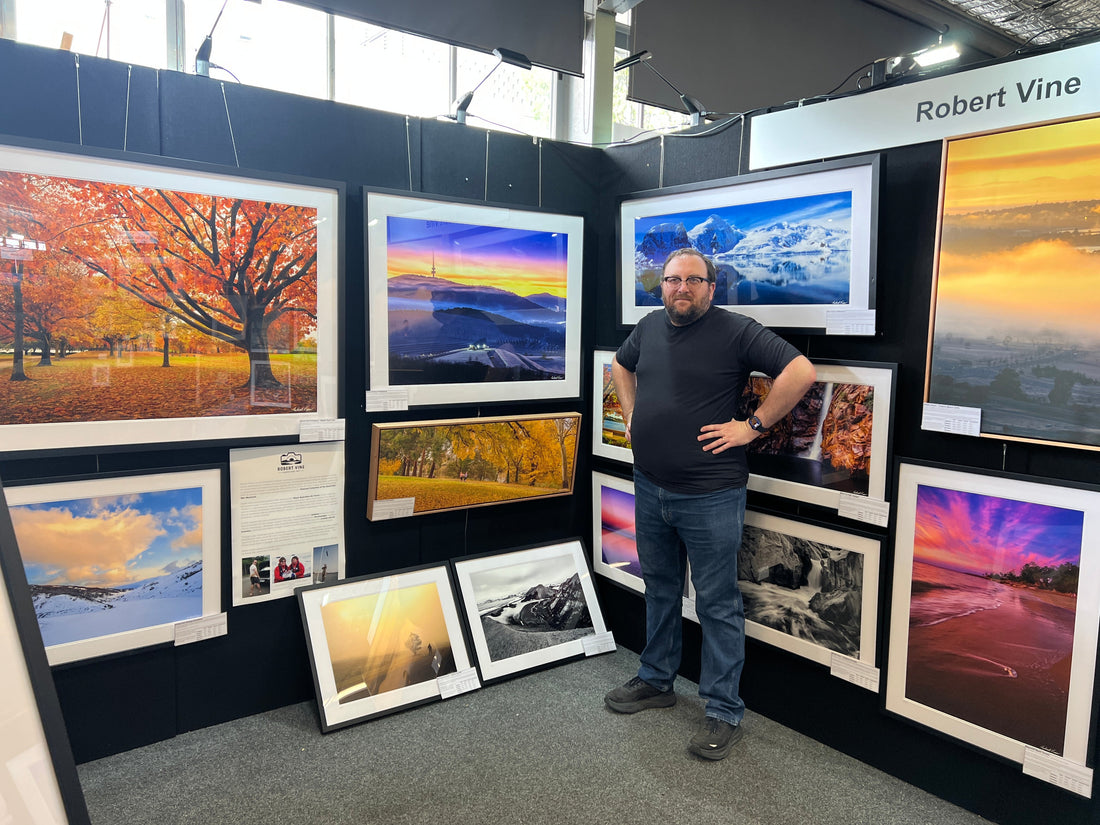 Robert Vine standing proudly at his Canberra Art Fair exhibition, surrounded by framed fine art landscape photographs featuring colourful Canberra autumn scenes, misty sunrises, Antarctic mountains and vibrant Australian landscapes.