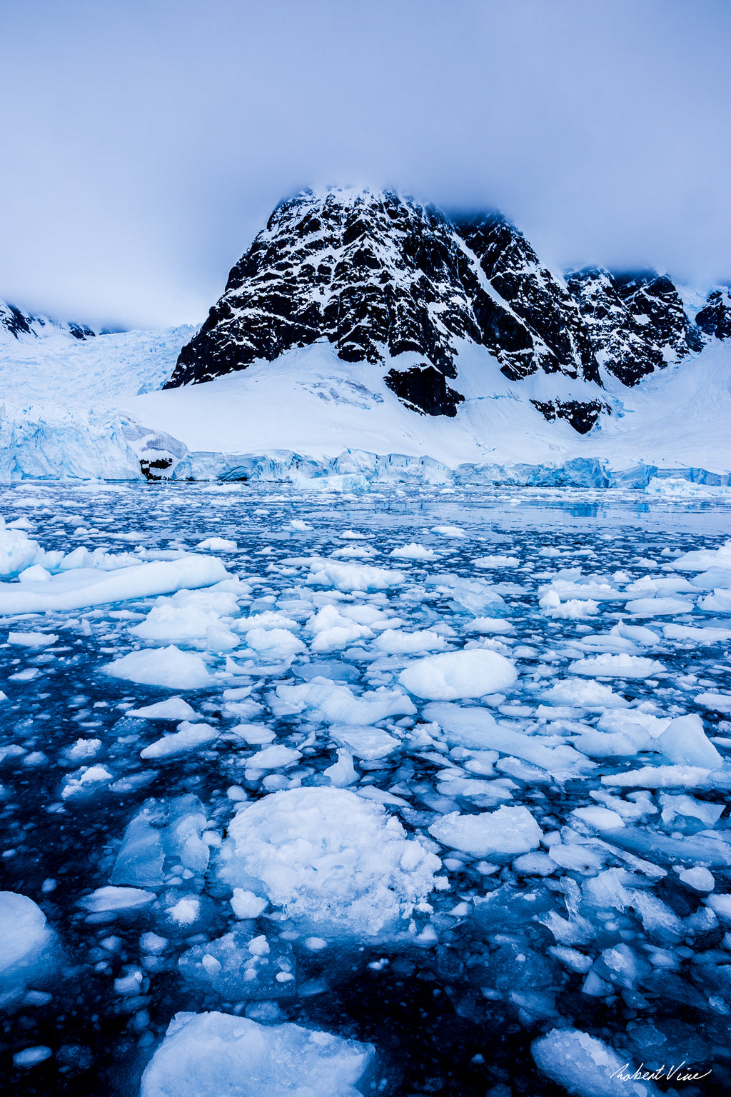 A dramatic Antarctic landscape at Paradise Bay featuring fractured sea ice, blue glacial water, and a snow-covered mountain rising into low cloud.