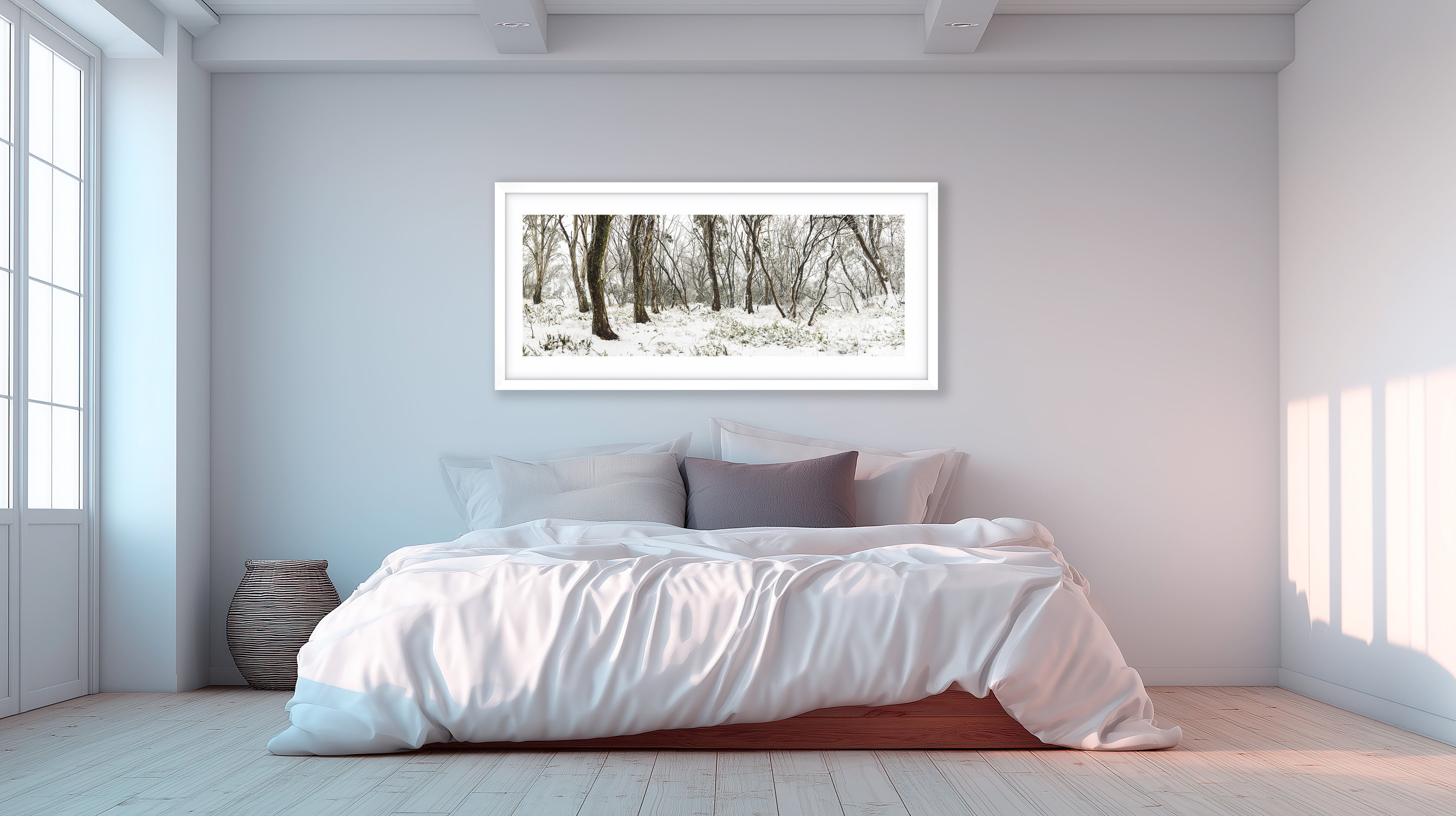 A serene minimalist bedroom with soft white bedding, pale timber floors, and a large framed panoramic winter forest art print on the wall above the bed, featuring snow-covered ground and bare trees. Light streams in from tall windows, creating a calm and modern atmosphere.