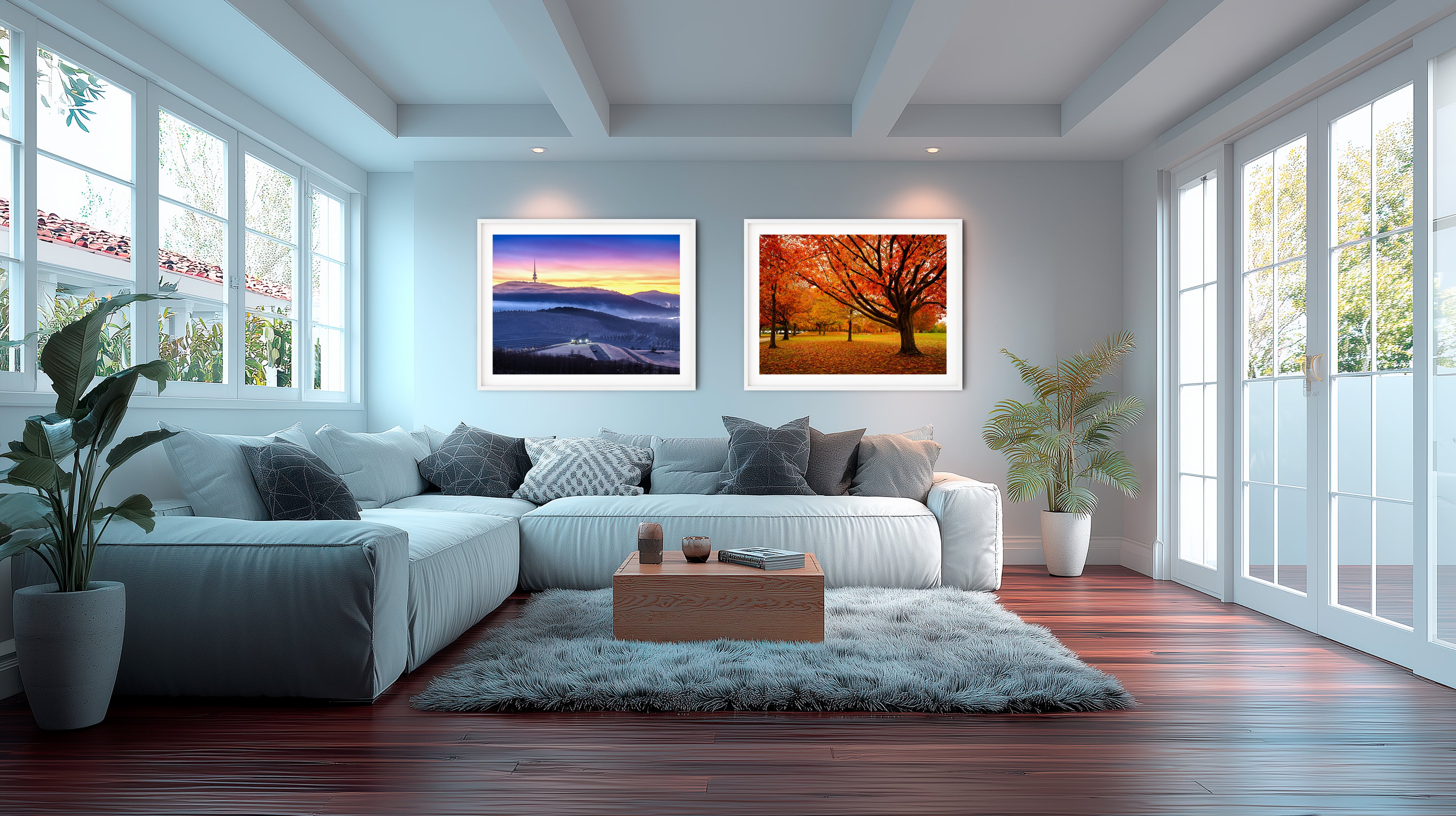 A bright modern Australian living room with a white sectional sofa, timber floorboards, large windows, indoor plants, and two framed art prints on the wall—one a vibrant sunrise over rolling hills and the other a colourful autumn tree scene.