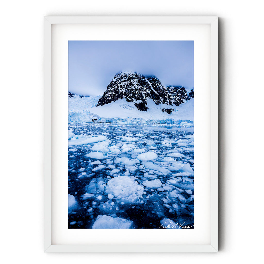A dramatic Antarctic landscape at Paradise Bay featuring fractured sea ice, blue glacial water, and a snow-covered mountain rising into low cloud.