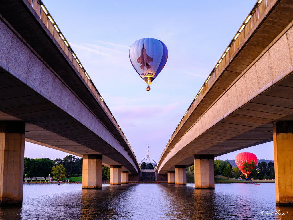 Air Force hot air balloon flying between Commonwealth Avenue Bridge spans with Parliament House in the background at dawn in Canberra.