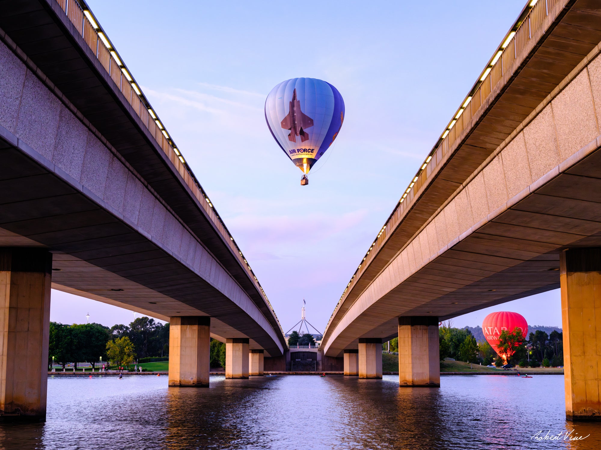 Air Force hot air balloon flying between Commonwealth Avenue Bridge spans with Parliament House in the background at dawn in Canberra.