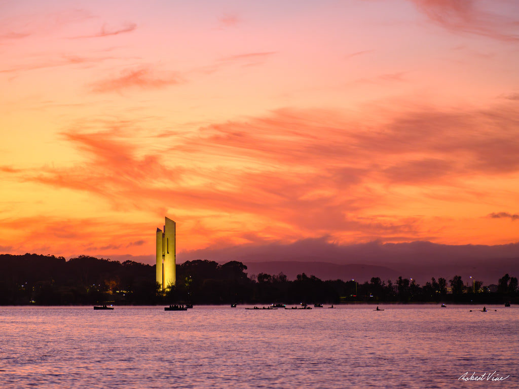 The National Carillon illuminated at sunrise on Lake Burley Griffin, Canberra, beneath vibrant orange and pink clouds reflected across calm morning waters.