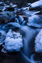 Long exposure photograph of the icy Thredbo River flowing between snow-covered granite boulders in the Australian Alps during winter.