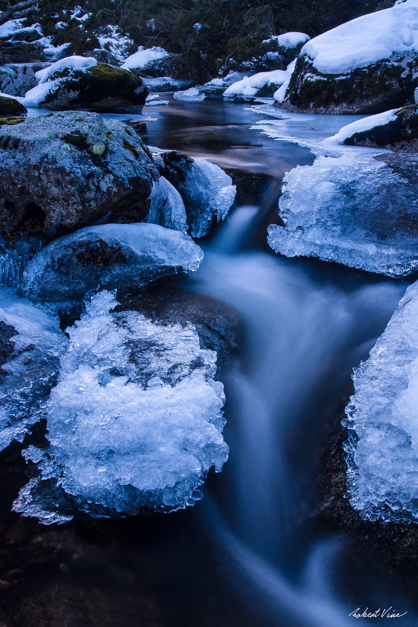 Long exposure photograph of the icy Thredbo River flowing between snow-covered granite boulders in the Australian Alps during winter.