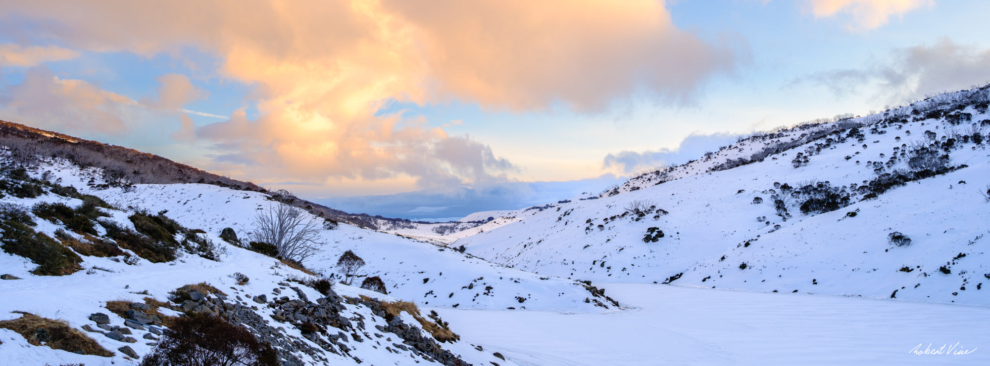 Panoramic winter sunset over the frozen Guthega Reservoir surrounded by snow-covered mountains in the Snowy Mountains, photographed by Robert Vine.