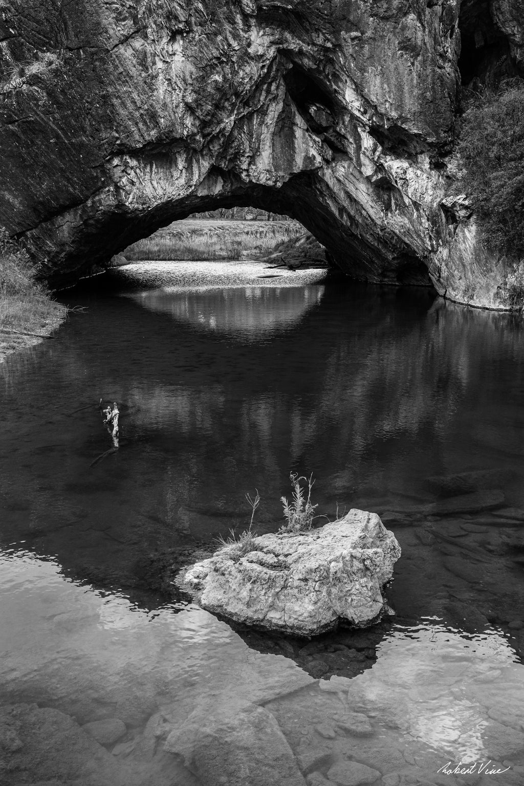 Black and white photograph of London Bridge at Googong, NSW, reflected in calm water beneath the natural limestone arch by Robert Vine.