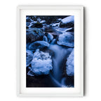 Long exposure photograph of the icy Thredbo River flowing between snow-covered granite boulders in the Australian Alps during winter.