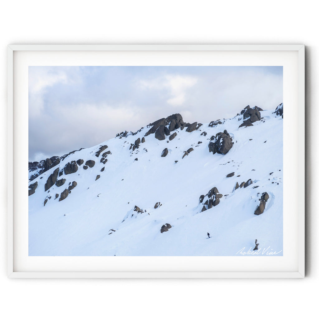 Framed wall art of a lone skier descending beneath snow-covered granite cliffs at Thredbo in the Snowy Mountains, photographed by Australian landscape artist Robert Vine.