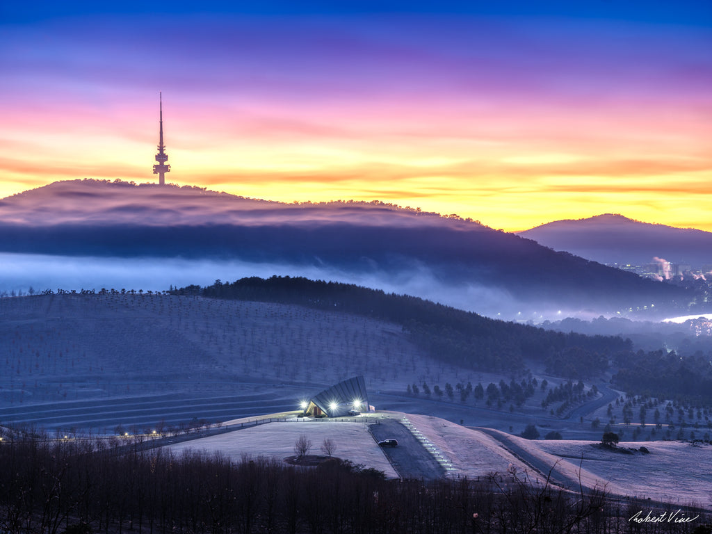 Frost-covered hills at the National Arboretum Canberra with Black Mountain Tower silhouetted against a colourful sunrise and layers of mist drifting through the valleys.